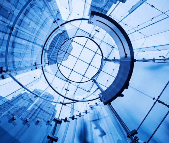 A modern glass spiral staircase viewed from below, with metal supports and a grid-patterned ceiling, surrounded by tall skyscrapers reflecting blue light.