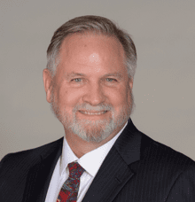 A middle-aged man with gray hair and a beard smiles, wearing a dark pinstripe suit, white shirt, and a patterned tie, posed against a plain light background.