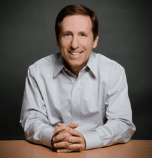A middle-aged man with brown hair, wearing a light blue button-up shirt, sits at a table with his hands folded and smiles at the camera against a dark background.