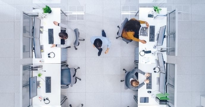 A top-down view of an office with four people working at separate desks with computers, and one person walking down the center aisle. The workspace is modern and organized with some green plants on desks.