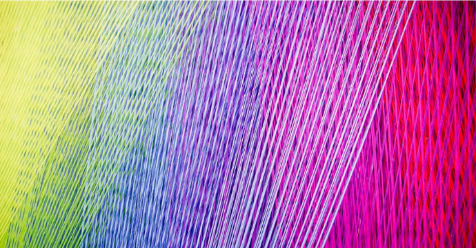Close-up of colorful threads arranged diagonally, transitioning from yellow on the left through green, blue, and purple, to pink and red on the right, creating a vibrant, textured pattern.