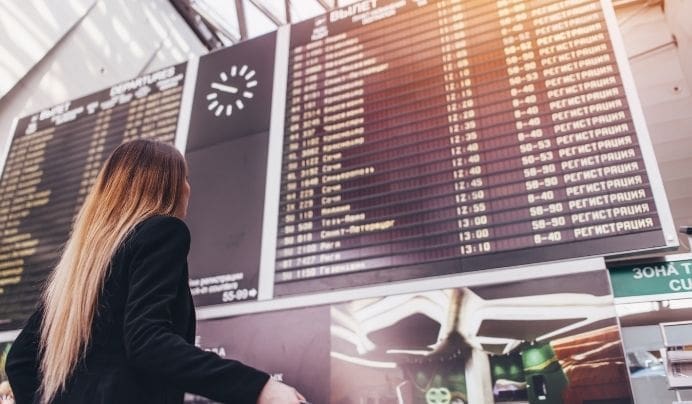 A woman with long, straight hair looks up at a large digital departure board inside an airport, checking flight times and information under a clock showing 11:40.
