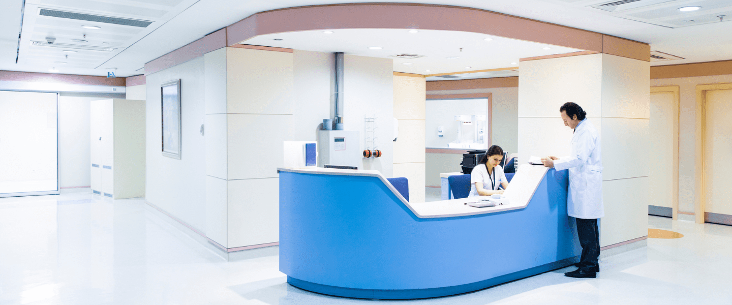 A doctor speaks to a receptionist who is working at a blue and white reception desk in a clean, modern hospital with bright lighting and minimal décor.