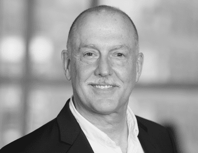 A smiling middle-aged man with a mustache and short hair, wearing a suit jacket over an open-collared shirt, stands in front of a blurred background. The image is in black and white.