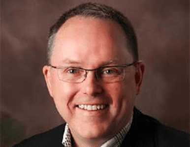 A smiling man wearing glasses, a dark jacket, and a light checked shirt poses in front of a brown, softly blurred background.