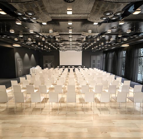 A modern conference room with rows of white chairs facing a large screen and podium at the front, wooden flooring, and a ceiling with exposed vents and circular lights.