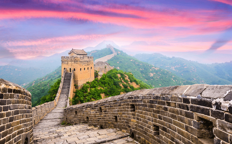 The Great Wall of China winds over green hills at sunrise, with a watchtower in the foreground and a colorful sky of pink and purple clouds overhead.