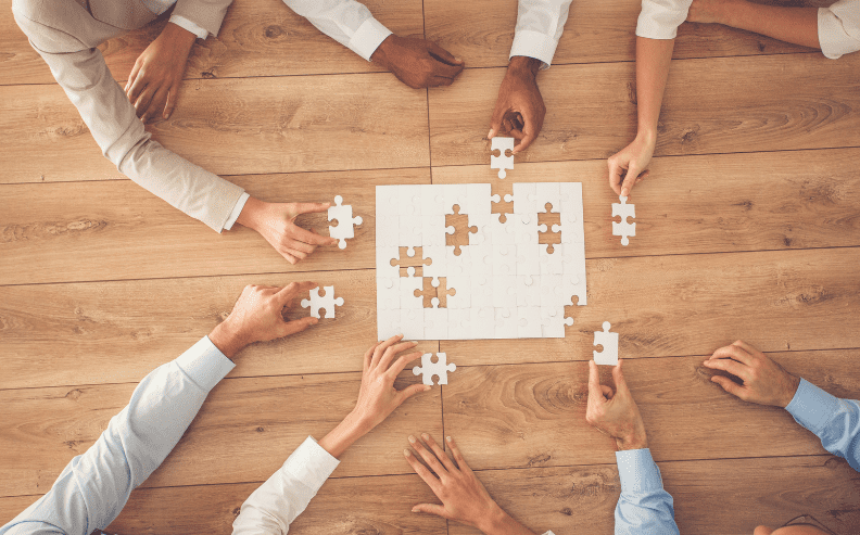Several people work together on a wooden table to complete a white jigsaw puzzle, each holding a puzzle piece and reaching toward the center where the puzzle is being assembled.