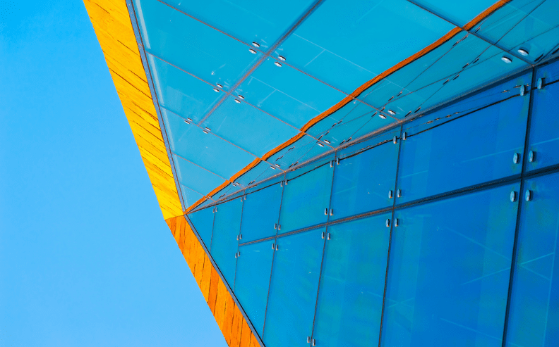 A close-up view of a modern building’s glass facade with blue-tinted windows and intersecting yellow structural elements, set against a clear blue sky.