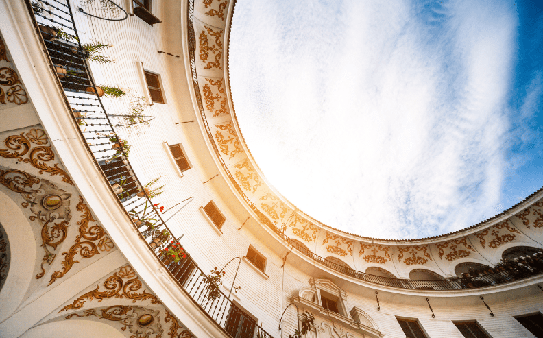 View looking up at a circular courtyard with ornate decorative patterns on the walls and balconies, plants hanging along the railings, and a bright, partly cloudy sky overhead.