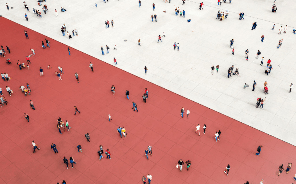 Aerial view of people walking and gathering on a large open plaza divided diagonally between red and white sections, creating a striking geometric contrast.
