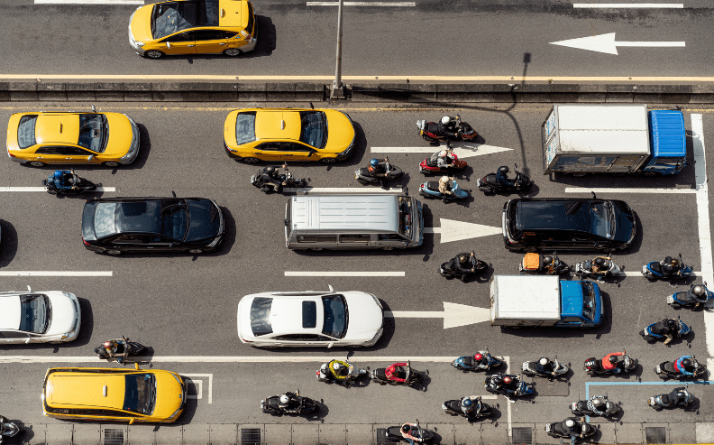 Aerial view of a busy city street with cars, taxis, trucks, and numerous motor scooters waiting in traffic at a stoplight, seen from above. White arrows are painted on the asphalt, indicating directions.