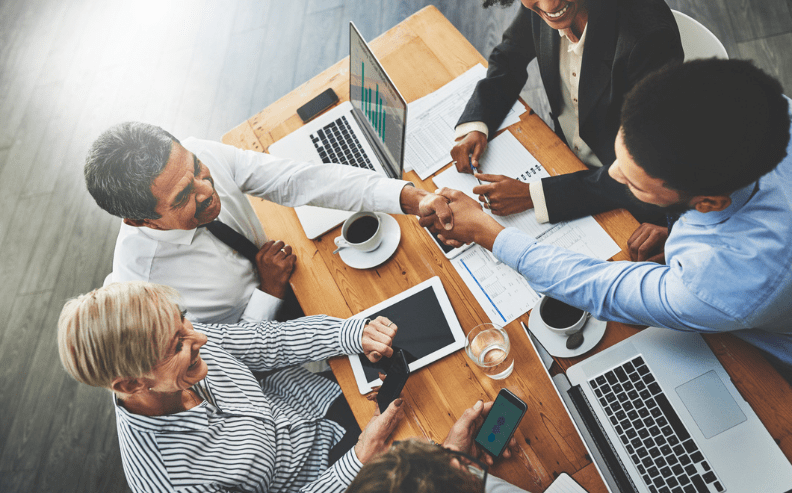 Four professionals sit around a wooden table with laptops, coffee, and documents, smiling and shaking hands, suggesting a successful business meeting or agreement.