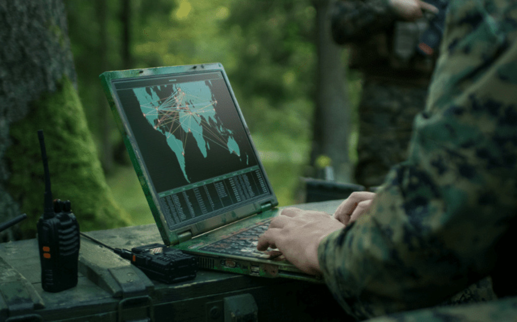 A person in camouflage uniform uses a rugged laptop displaying a world map with network connections. A two-way radio sits nearby, and the scene appears to be outdoors in a wooded area.