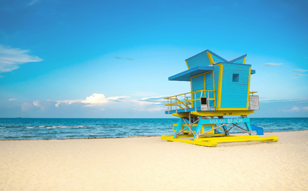 A bright blue and yellow lifeguard tower stands on the sandy beach near the ocean under a clear blue sky, with gentle waves and clouds visible in the distance.