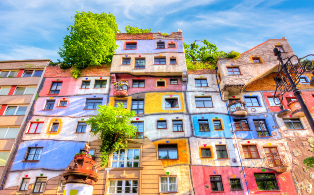 Colorful, irregularly shaped apartment building with painted facades, uneven windows, and balconies, covered in vibrant hues and accented by greenery, against a bright blue sky.