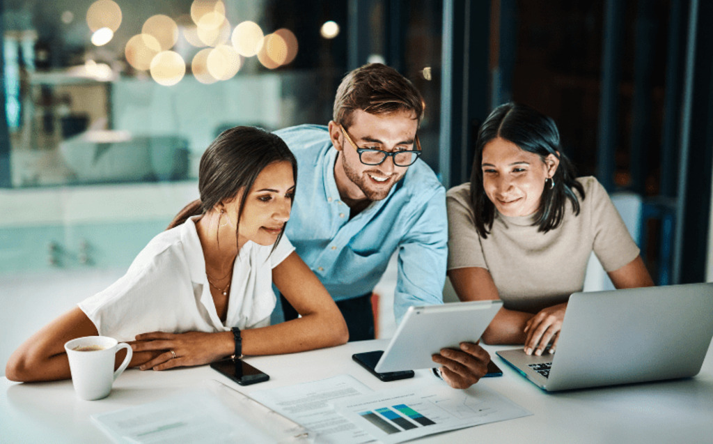 Three colleagues sit at a desk in an office at night, looking at a tablet and laptop. Papers with charts are on the table, and city lights are visible through large windows in the background.