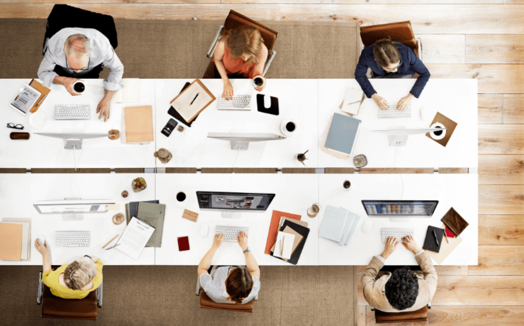 Six people work at a shared office table, viewed from above. Each person uses a computer and has papers, notebooks, and coffee cups on their desks. The workspace is modern with light wood floors.
