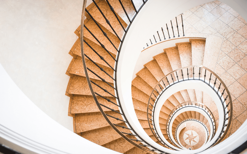 A top-down view of a spiral staircase with light brown steps and black railings, creating a circular, geometric pattern as it descends to a tiled floor below.