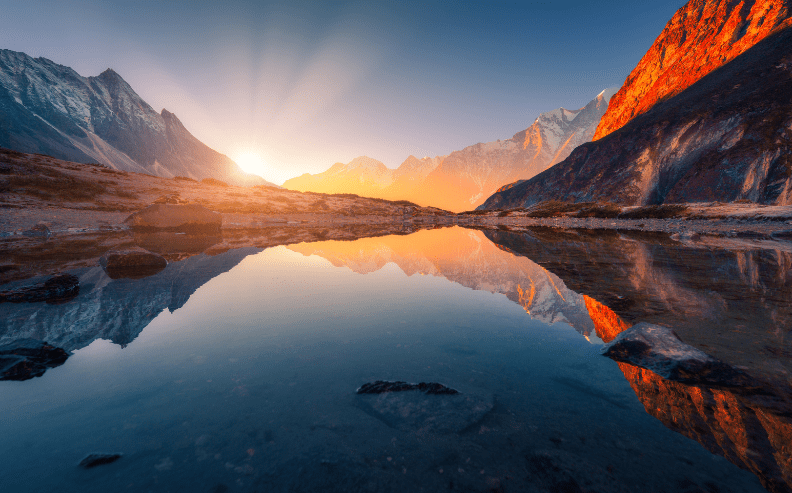 Sunrise over a mountain range with sunlight streaming across snow-capped peaks, reflected in a calm lake. Rocky shores and vibrant orange light illuminate the landscape.
