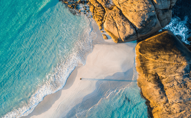 Aerial view of turquoise waves washing onto a sandy beach bordered by large, sunlit rocky formations, with a single person walking and casting a long shadow on the shore.