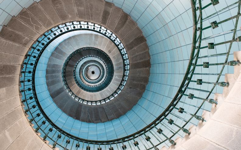 View looking up a spiral staircase with blue-tiled walls and green railings, creating a circular, hypnotic pattern that draws the eye toward the center at the top.