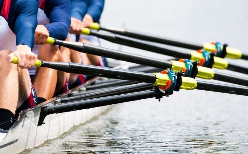 Close-up of a rowing team in action, showing synchronized arms and oars moving through the water, creating small ripples. The rowers wear matching uniforms with blue sleeves.