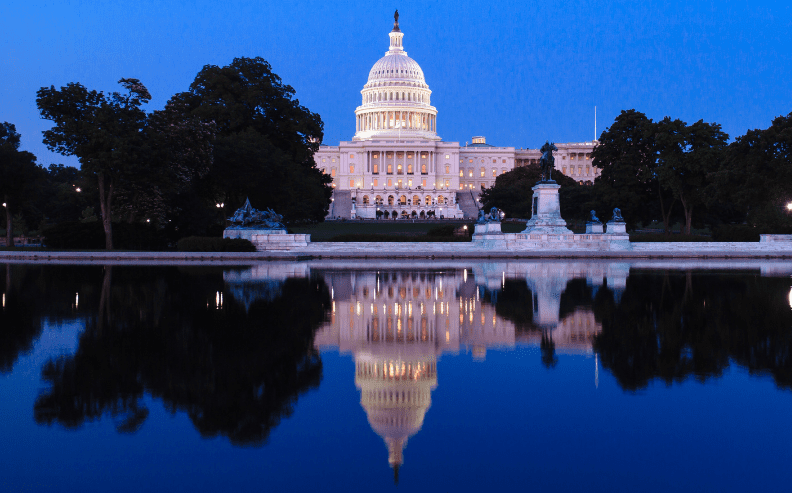 The U.S. Capitol building at dusk, illuminated with lights, is reflected in a calm body of water. Trees and statues surround the building, and the sky is deep blue.