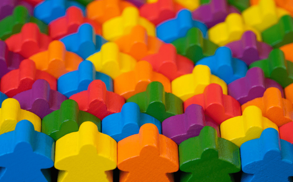 A close-up of many colorful wooden game pieces, shaped like people, arranged in a dense, overlapping pattern. The pieces are in bright colors such as red, blue, green, yellow, orange, and purple.