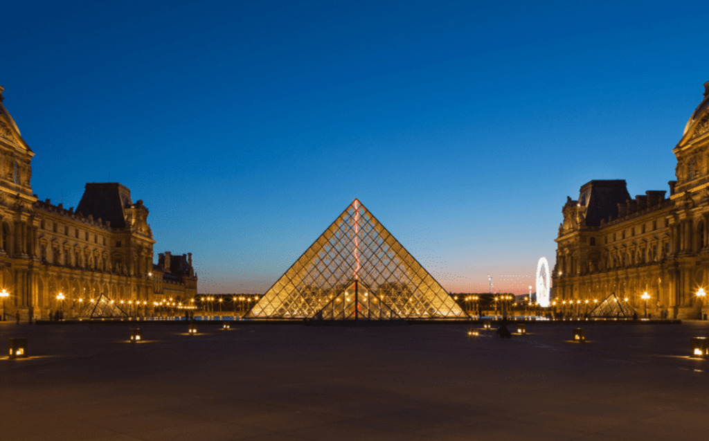 The Louvre Museum in Paris at dusk, with the glass pyramid entrance brightly illuminated in the center, flanked by historic buildings and a deep blue sky above.