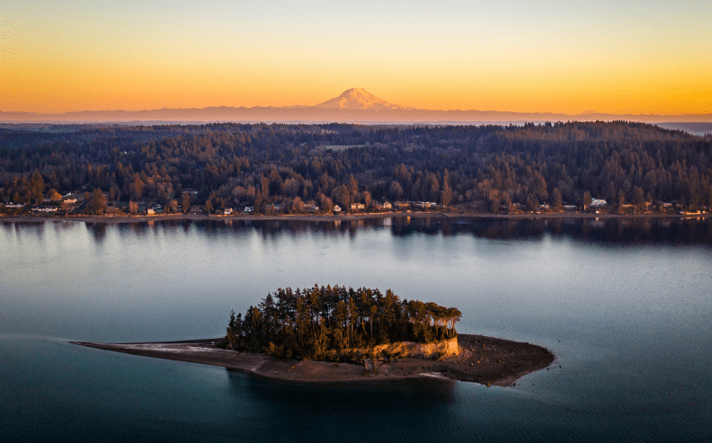 A small, tree-covered island sits in calm water at sunset, with a forested shoreline and scattered houses in the background. A snow-capped mountain rises in the distance under a golden sky.