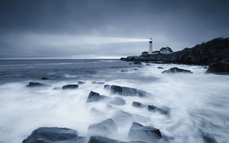 A lighthouse shines its beam across a rocky coastline under a cloudy, dark blue sky, with waves crashing against the rocks in the foreground.