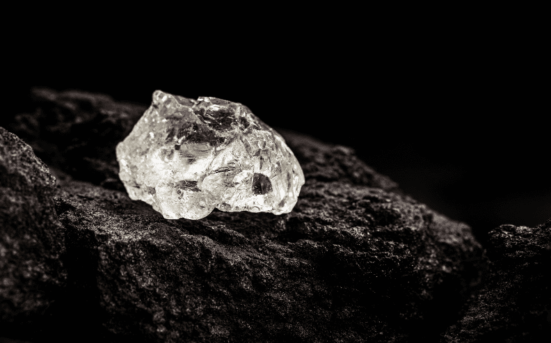 A rough, uncut diamond sits on top of a dark, textured rock with a black background.