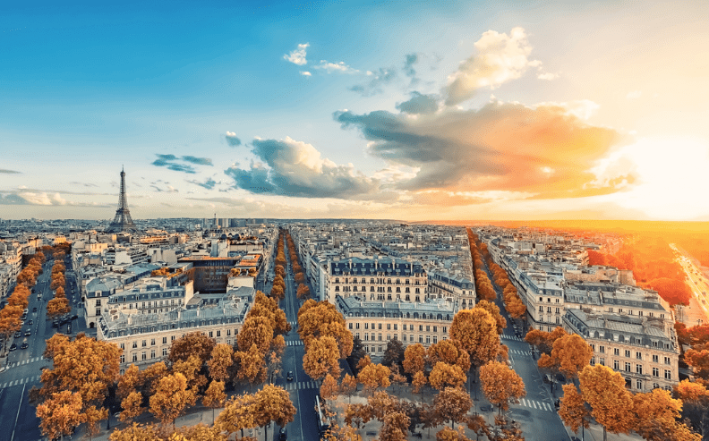 A wide aerial view of Paris at sunset, showing tree-lined streets, classic buildings, and the Eiffel Tower in the distance beneath a partly cloudy sky with bright sunlight.