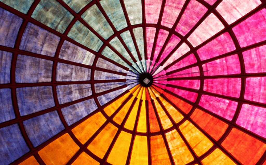 A colorful stained glass ceiling features radiating sections in shades of red, pink, blue, orange, and yellow, arranged in a circular pattern with dark grid lines dividing each segment.