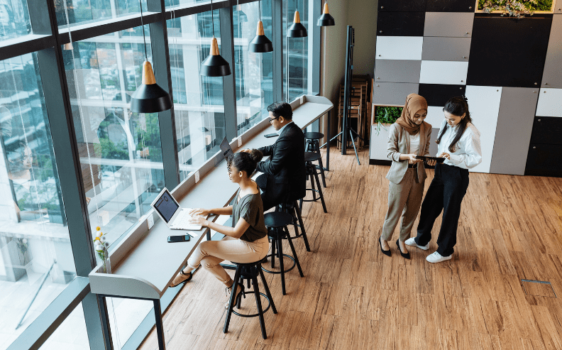 Four people work in a modern office. Two sit at high desks by large windows using laptops, while two women stand together on the right, looking at a tablet and discussing something. The room has wood floors and hanging lights.
