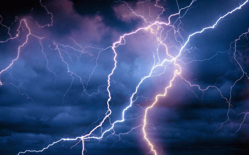 Bright lightning bolts illuminate a dark, cloudy night sky, creating dramatic zigzag patterns and highlighting the deep blue and purple hues of the storm clouds.