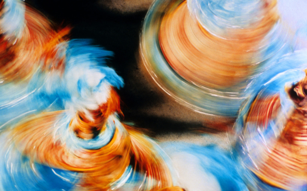 Abstract image showing blurred, swirling shapes in orange, blue, and white, suggesting spinning dancers in motion with colorful flowing skirts and dynamic movement against a dark background.