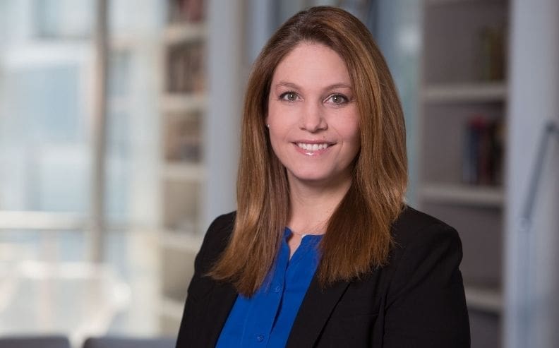 A woman with straight, light brown hair wearing a blue blouse and black blazer smiles at the camera in a modern office setting with shelves and large windows in the background.