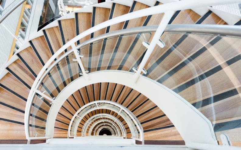 A spiral staircase with wood-patterned steps and a white railing, viewed from above, creates a circular, hypnotic pattern as it descends several floors.