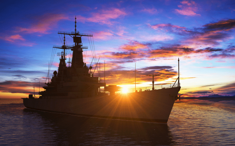 A large navy battleship is silhouetted against a vibrant sunset, with colorful clouds and the sun low on the horizon, reflecting on the calm ocean water.