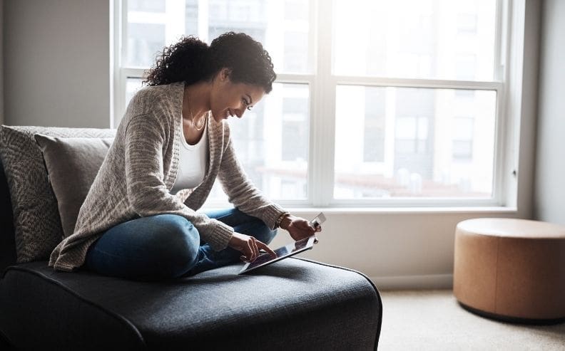 A woman sits cross-legged on a couch near a large window, using a tablet. She is holding a credit card and appears focused, with sunlight streaming into the modern room.