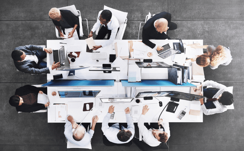 A group of professionals sit around a large conference table, working on laptops, reading papers, and discussing, viewed from above in a modern office setting.