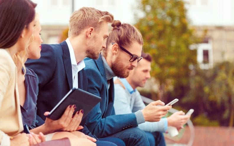 Four young professionals sit outdoors in a row, dressed in business attire. Two are looking at mobile devices, one holds a tablet, and they appear engaged with their technology, with trees and blurred buildings in the background.