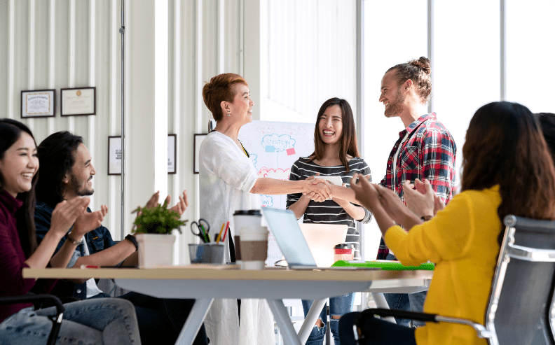 A group of people in a modern office celebrate as two colleagues stand and shake hands, smiling, while others sit around a table clapping and cheering.