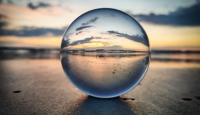 A clear glass sphere on wet sand reflects a colorful sunset sky with clouds, creating an inverted, sharp image of the beach inside the ball; the background is softly blurred.