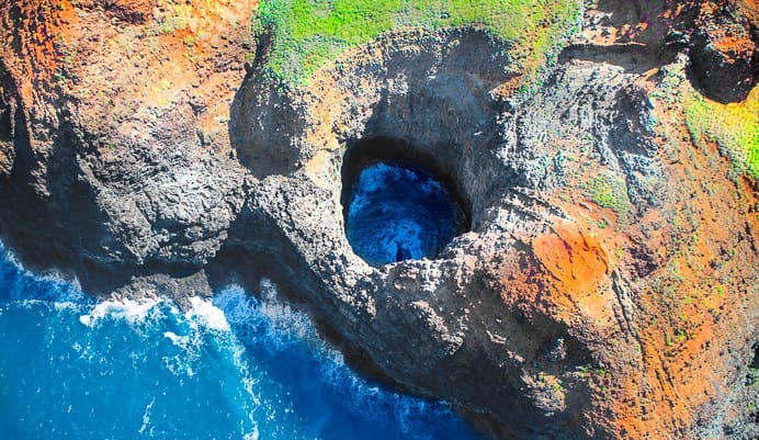 Aerial view of a rocky coastal cliff with a large circular hole through which blue ocean water is visible, surrounded by green and orange vegetation.
