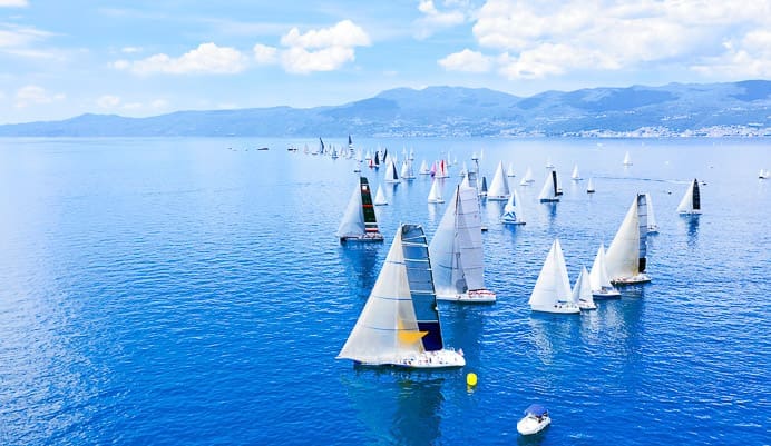A large group of sailboats with white sails participates in a regatta on calm blue water with distant mountains and a partly cloudy sky in the background. A small motorboat is near the sailboats.