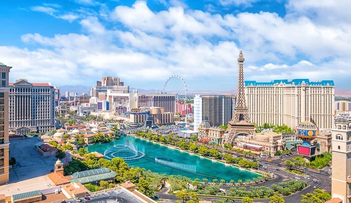 Aerial view of the Las Vegas Strip featuring famous casinos, hotels, a large fountain, and a replica of the Eiffel Tower under a partly cloudy blue sky.