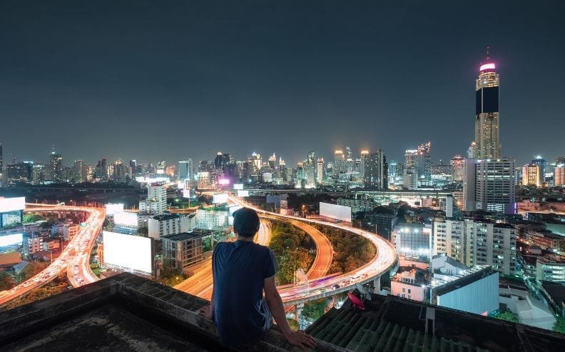 A person sits on a rooftop at night, overlooking a brightly lit city skyline with tall buildings and winding highways filled with moving car lights.